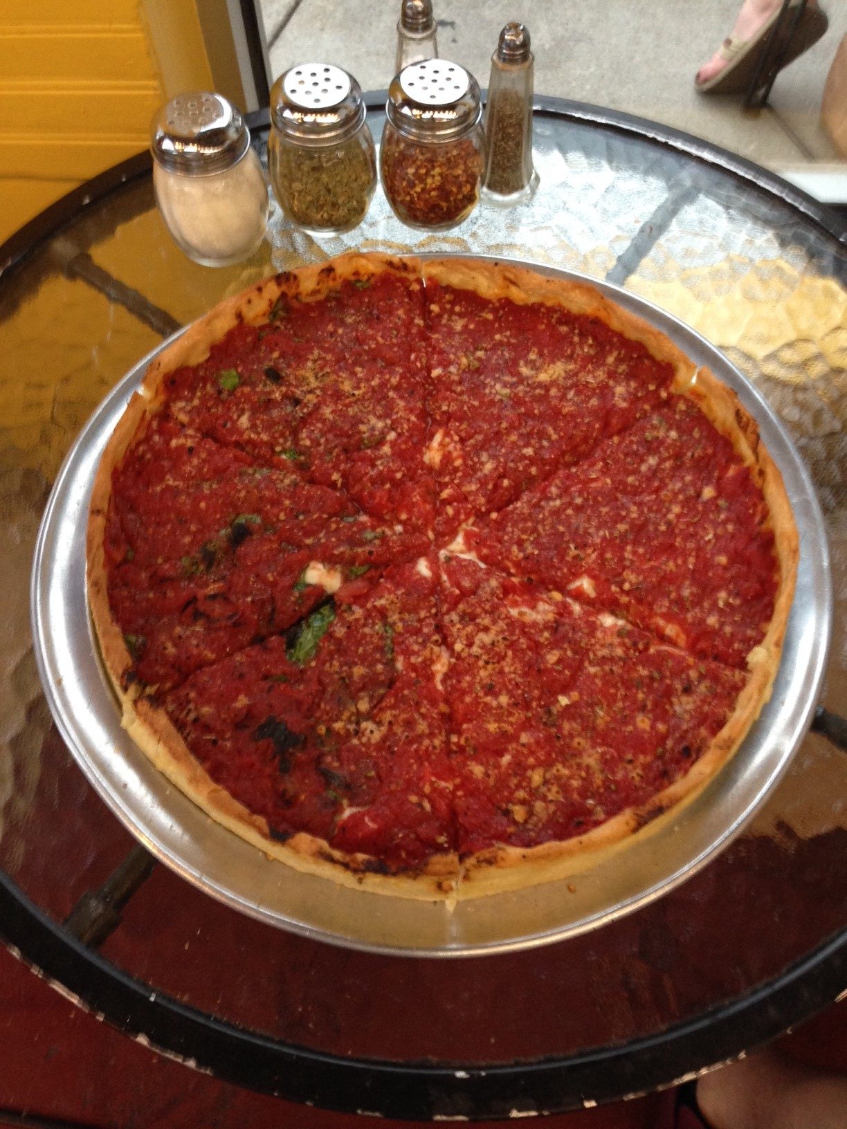 A deep dish pizza on a metal pan, with spice shakers on the table